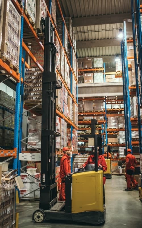 Person driving a skid loader with vertical shelfing and people moving products around a warehouse